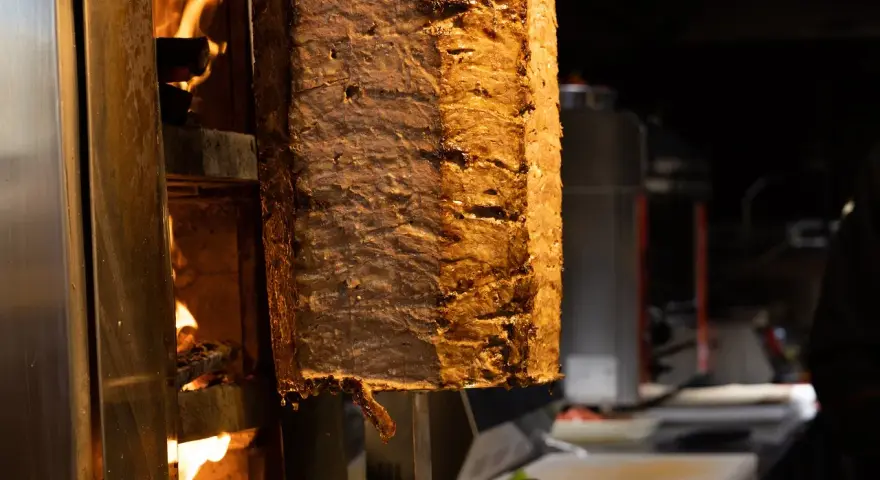 Duży pionowy obrotowy grill z mięsem, obok płomienie ognia.