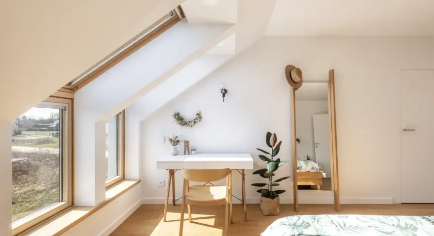 Angolo studio mansardato inondato da la luce più naturale. Scrivania, sedia in legno, specchio alto e pianta.