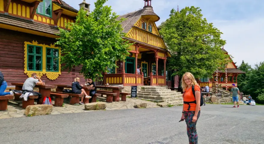 Kobieta na tle kolorowych, drewnianych budynków w stylu pustevny. Ludzie siedzą przy stołach na zewnątrz.
