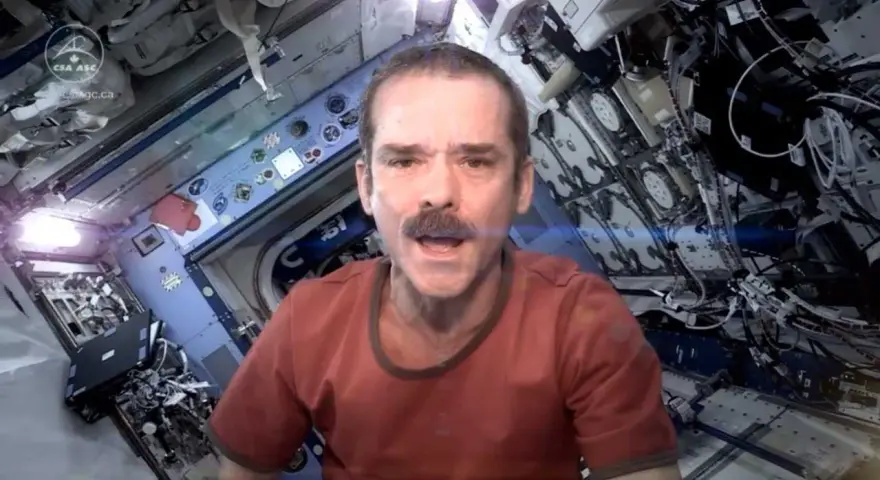 Chris Hadfield singt "Space Oddity" in der ISS. Sein Gesicht mit Schnurrbart ist im Vordergrund, der Raumschiff-Innenraum im Hintergrund.