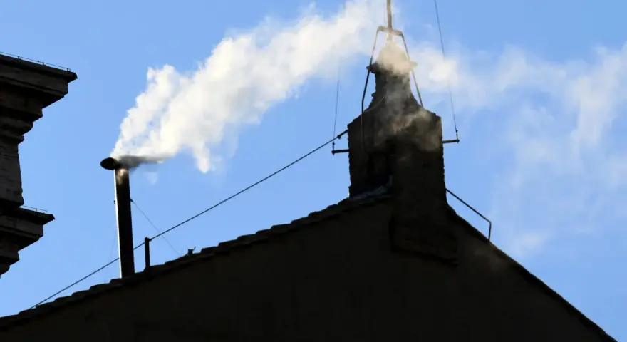 Biały dym z komina, jak sygnał wyboru nowego papieża, unosi się na tle błękitnego nieba.