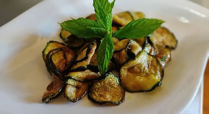 Zucchine alla scapece dorate e croccanti, guarnite con foglie di menta fresca, servite su un piatto bianco.