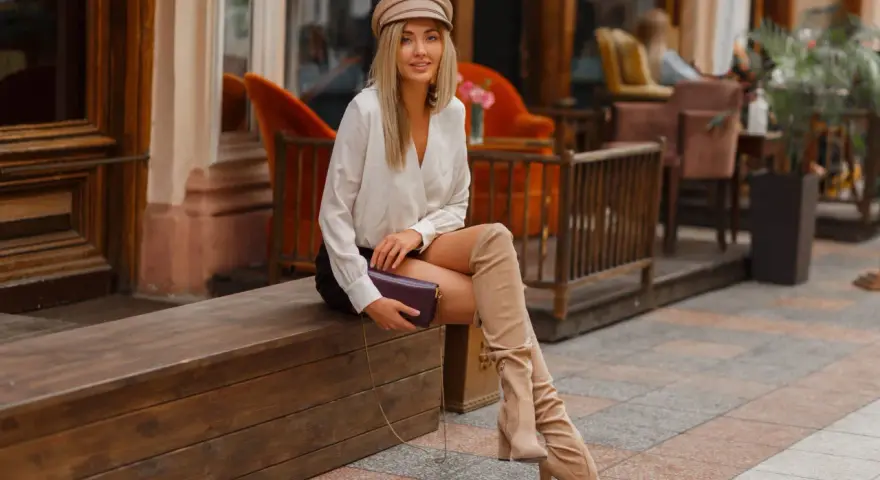 Une femme élégante assise dehors, portant une tenue chic avec des bottes hautes beiges. Idée de comment porter des bottes hautes marrons avec style.