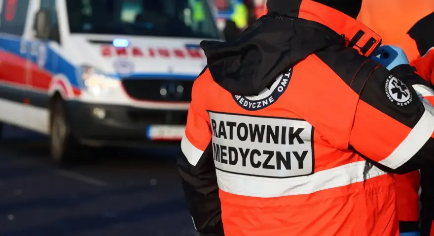 Ratownik medyczny w jaskrawej kurtce stoi obok karetki pogotowia.