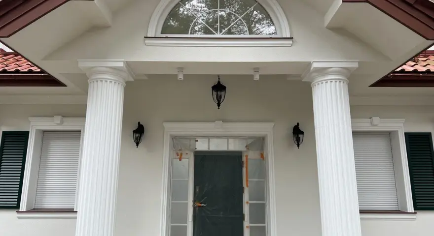 Elegancki pilaster zdobi fasadę domu, wspierając dach nad wejściem. Obok okna i drzwi, tworzy klasyczną kompozycję.