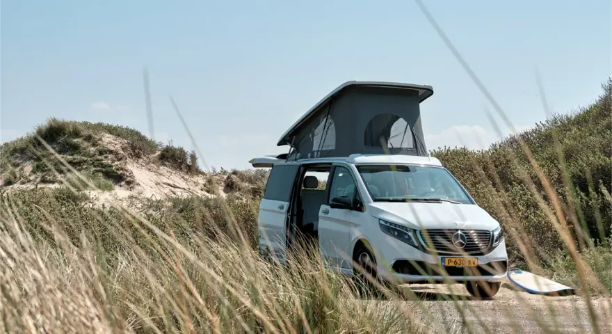 Vit el campervan med uppfällt tak står parkerad vid en sanddyn. En surfbräda ligger bredvid.