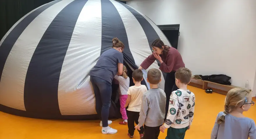 Dzieci i opiekunki wchodzą do dmuchanej kopuły, która przypomina planetarium. To miejsce, gdzie można oglądać filmy z przesłaniem, odkrywając tajemnice wszechświata.