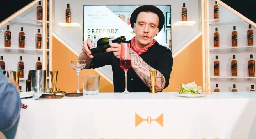 Barman Grzegorz Pikuła, w stylowym kapeluszu, nalewa drinka. W tle półki z butelkami whisky, idealne dla miłośników krwawych mary.