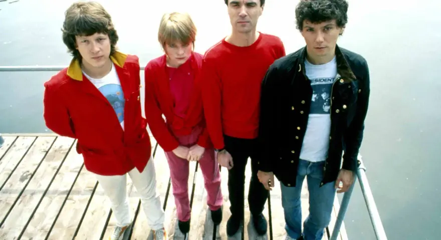 Die Band Talking Heads steht auf einem Holzsteg. David Byrne trägt ein rotes Hemd, Tina Weymouth ein pinkes Outfit.