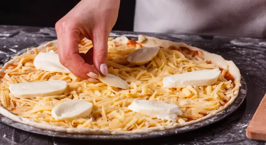 Dłoń układa kawałki sera na pizzy, która jest już posypana startym żółtym serem.