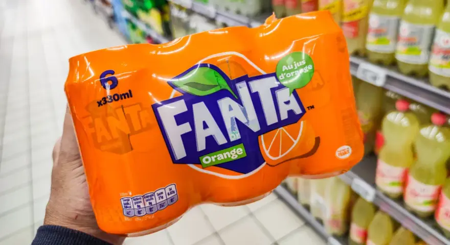 Opakowanie 6 puszek Fanty Orange w ręce, na tle sklepowych półek z napojami.