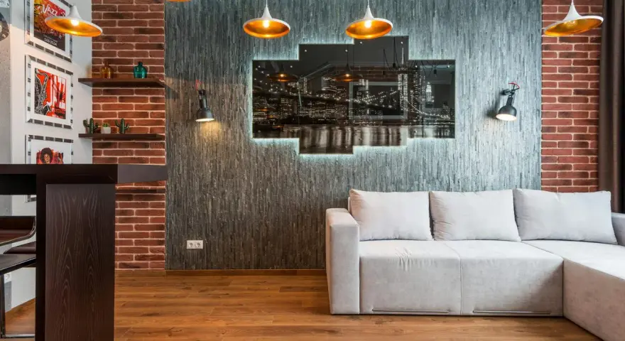 Nowoczesny salon w stylu loft: szara kanapa, ceglana ściana, panel z nocnym widokiem miasta i złote lampy.