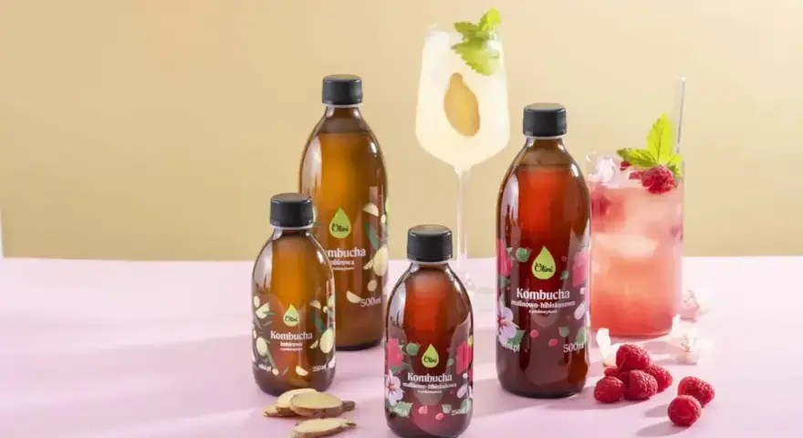 Butelki kombuchy o różnych smakach: imbirowej i malinowo-hibiskusowej. Obok napojów dwa drinki, maliny i plasterki imbiru. Poczuj, jak smakuje kombucha!