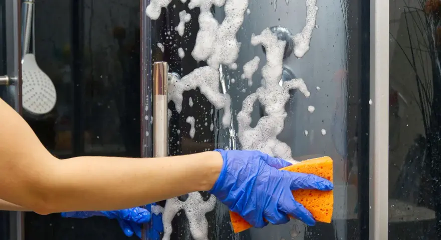 Main bleue, éponge orange pour nettoyer paroi douche. Mousse et bulles sur la paroi vitrée.