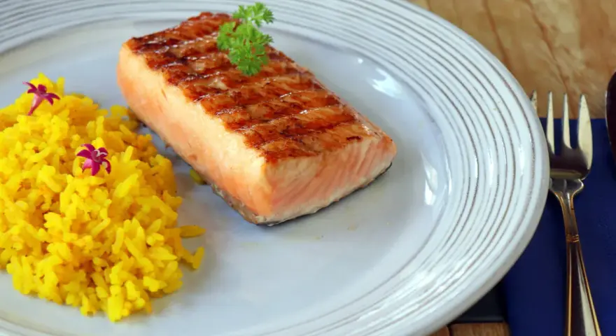 Un delicioso salmón a la plancha con marcas de parrilla, servido con arroz amarillo y decorado con perejil y flores moradas.