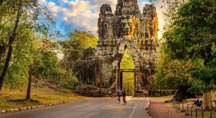Two cyclists approach a massive stone gateway, one of the ancient Cambodia temples, bathed in golden sunlight.