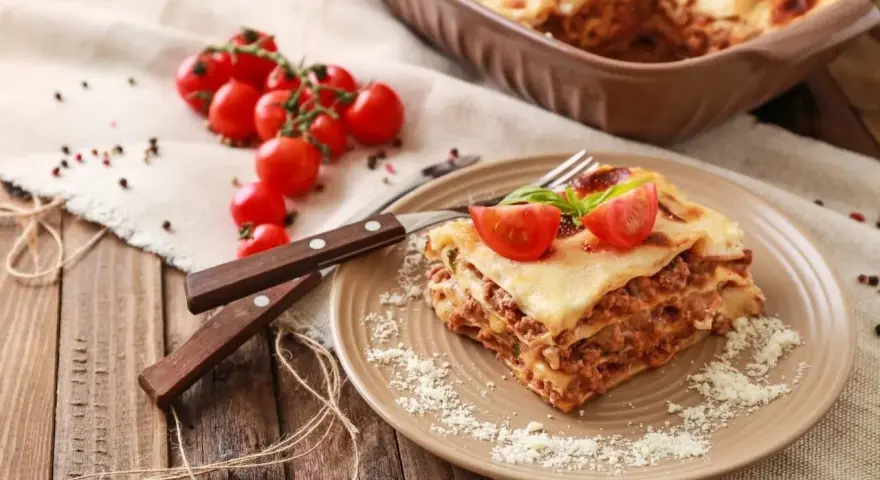 Pyszna, wielowarstwowa lasagne z mięsem mielonym, polana sosem beszamelowym i posypana parmezanem. Na talerzu ozdobiona pomidorkami koktajlowymi i bazylią.