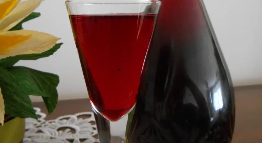 Szklanka z nalewką z aronii mrożonej na wódce, obok ciemna butelka. W tle ozdobny kwiat.