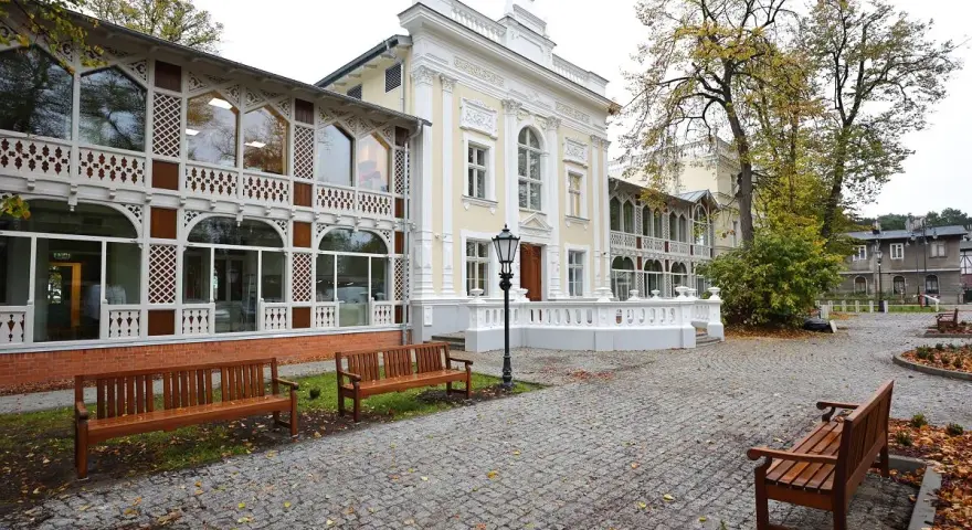 Elegancki budynek z ażurowymi werandami, przypominający dawny **Hevelianum dom zdrojowy**. Brukowany dziedziniec z ławkami i jesiennymi liśćmi.