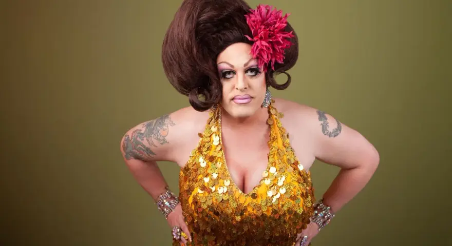 Osoba w złotej, cekinowej sukience i peruce, z kwiatem we włosach. To przykład tego, co to jest drag queen – artystyczna forma ekspresji.
