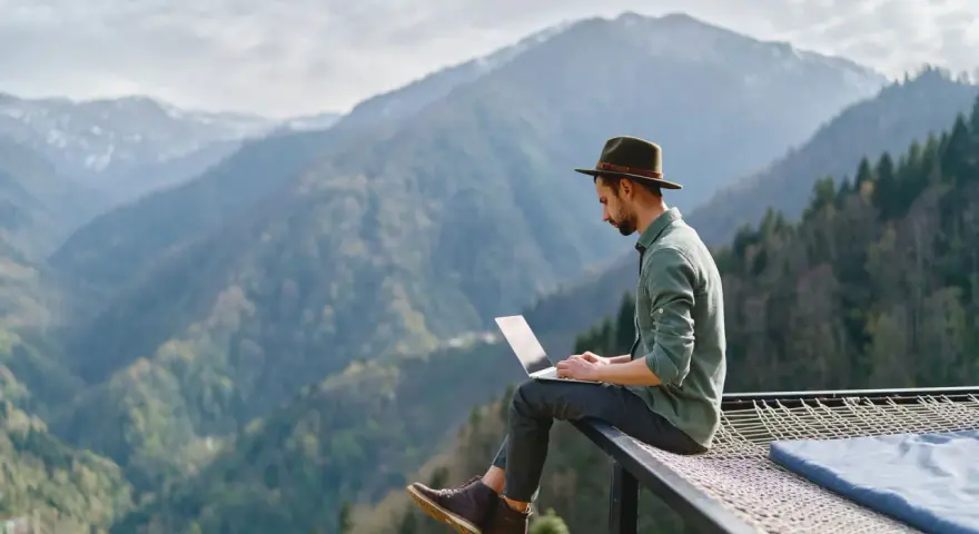 Un homme en chapeau travaille sur son ordinateur portable, profitant du télétravail à l'étranger avec une vue imprenable sur les montagnes.