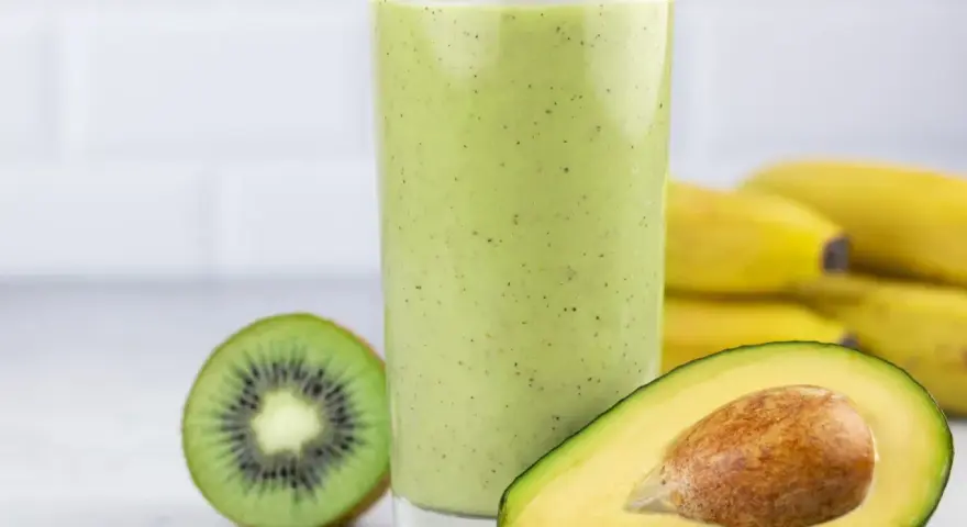 Zielony koktajl z awokado, kiwi i bananów w szklance, obok połówka awokado i kiwi.