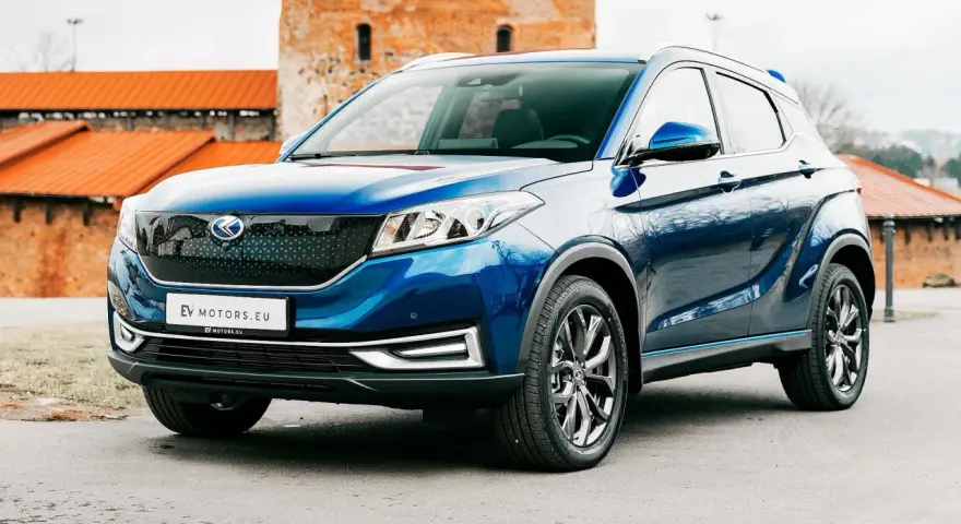 Błękitny SUV EV Motors.eu z charakterystycznym grillem, zaparkowany przed zabytkowym budynkiem z cegły.