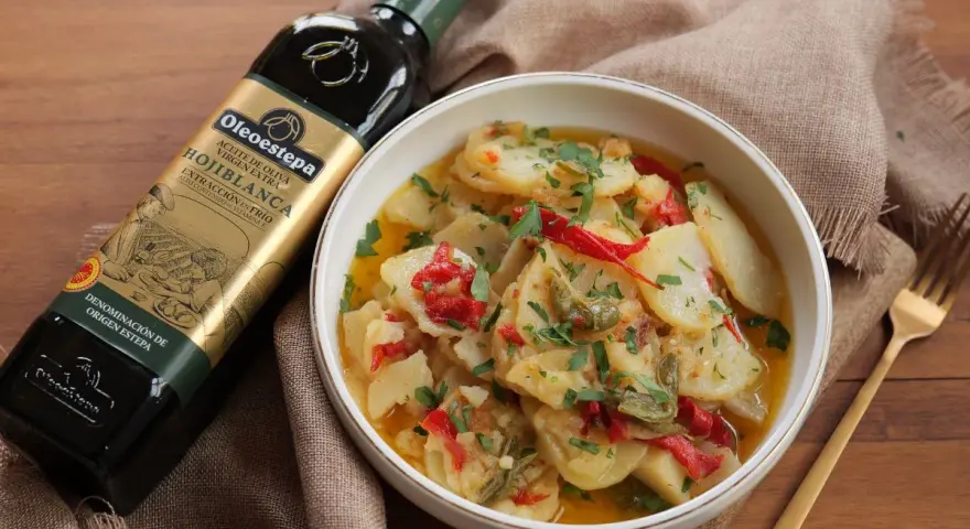 Un tazón de patatas a lo pobre, con pimientos rojos y verdes, perejil picado y una botella de aceite de oliva virgen extra.