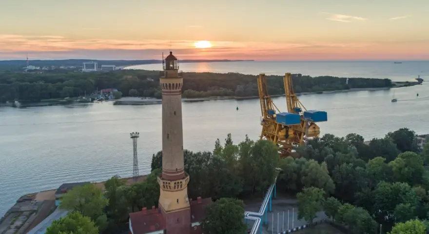 Zachód słońca nad morzem, obok najwyższej latarni morskiej w Polsce, widać też portowe dźwigi.
