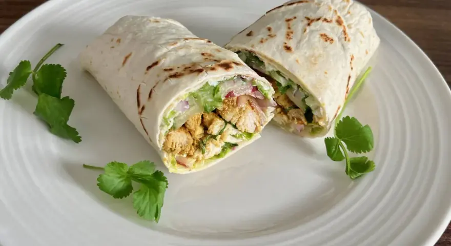 Kebab w tortilli przekrojony na pół, z kurczakiem, warzywami i kolendrą na białym talerzu.
