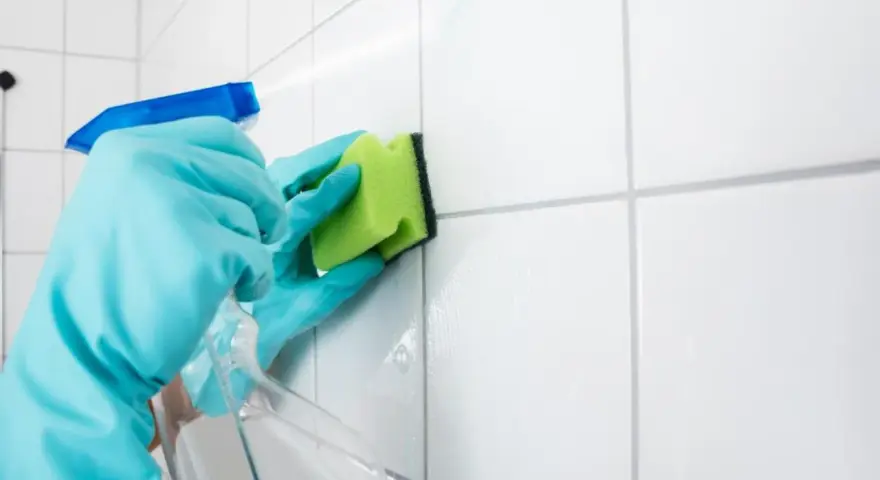 Manos con guantes azules rociando limpiador y frotando juntas de azulejos blancos con una esponja verde para blanquear juntas azulejos.