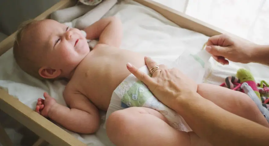 Radość noworodka podczas pielęgnacji. Mama zmienia pieluszkę, maluch uśmiecha się, leżąc na przewijaku wśród zabawek.