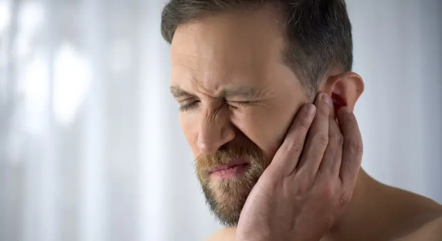 Mężczyzna z zarostem cierpi z powodu bólu chrząstki ucha przy dotyku, zaciskając oczy z bólu.