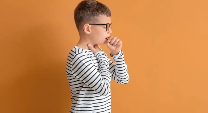 Chłopiec w okularach ma **szczekający kaszel u dziecka**. Dotyka gardła, czując dyskomfort.