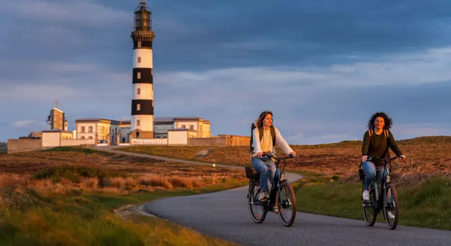 Deux femmes font du vélo sur une route sinueuse à Ouessant, avec un phare emblématique en arrière-plan. Une belle façon de visiter Ouessant à pied, ou plutôt à vélo !