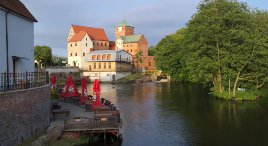 Zamek Darłowo z wieżami i zielonymi dachami, otoczony wodą i drzewami. Przy brzegu letnia restauracja z czerwonymi parasolami.