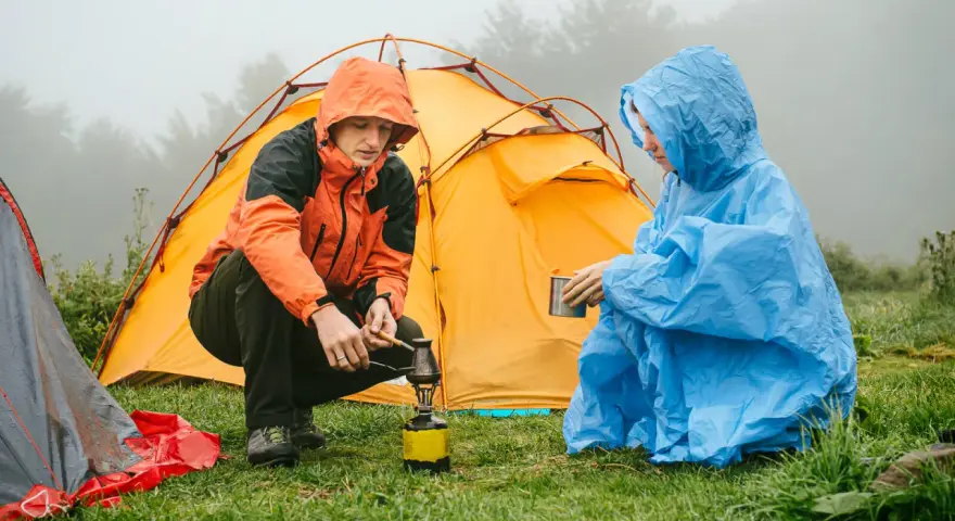 Zwei Camper in Regenponchos bereiten sich auf das Zelten bei Regen vor. Einer kocht auf einem Campingkocher, der andere hält eine Tasse.