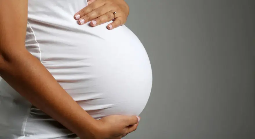 Brzuch ciężarnej kobiety w białej bluzce, dłonie delikatnie go obejmują. Zbliża się poród, może już wkrótce pojawi się czop śluzowy.