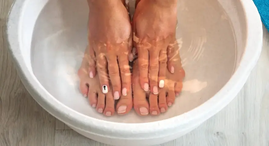 A relaxing home remedy foot soak in a white basin. Feet with french manicures and one toe nail decorated with a black and white design are submerged in clear water.