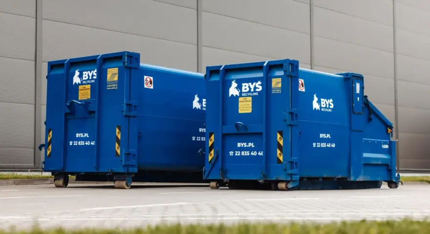 Dwa niebieskie kontenery BYS Recykling stoją przed budynkiem. Dowiedz się, ile kosztuje tona gruzu betonowego i jak go zamówić.