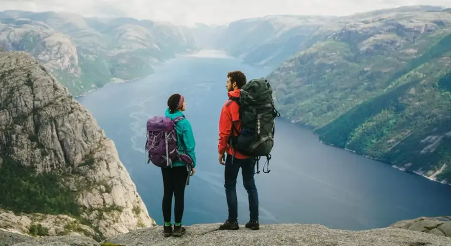 Två vandrare med stora ryggsäckar står på en klippa och blickar ut över en fjord i Norge. Perfekt för camping i Norge.
