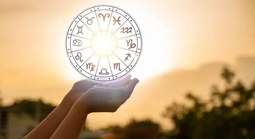 Dłonie trzymają koło zodiaku, gdzie widoczny jest symbol Wagi. Słońce przebija się przez niebo, tworząc magiczną aurę.