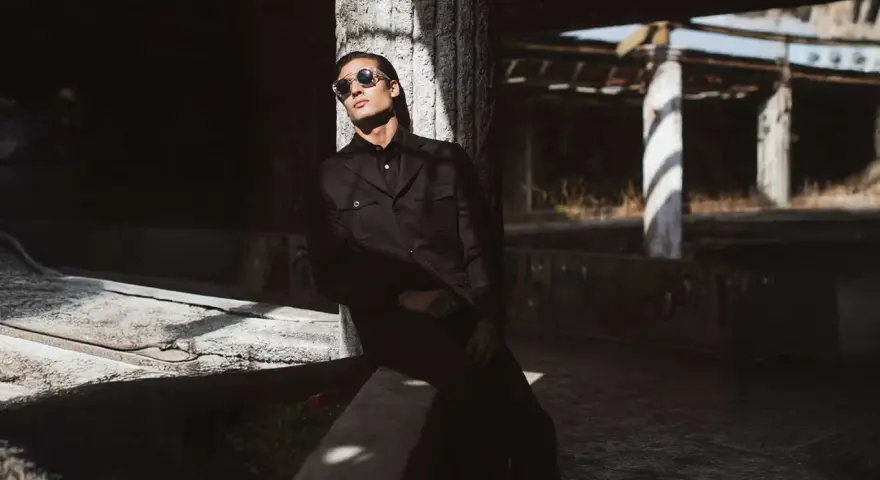 Hombre con gafas de sol y un **smart outfit** oscuro, apoyado en un pilar de hormigón en un entorno urbano abandonado.