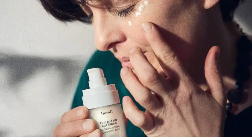 Twarz 50 latki z aplikacją kremu pod oczy. Dłonie delikatnie rozprowadzają produkt, dbając o skórę.