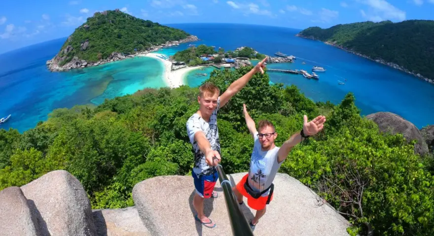 Dwóch mężczyzn robi selfie na tle tropikalnej wyspy z turkusową wodą i plażą.