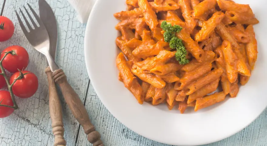 Piatto di penne alla vodka cremose, guarnite con prezzemolo, su un tavolo di legno azzurro. Accanto, pomodorini freschi e posate rustiche.