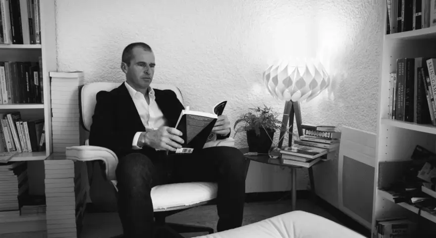 Un homme lit un livre dans un fauteuil, entouré de livres. La lumière d'une lampe éclaire la scène. ae-l af-l.