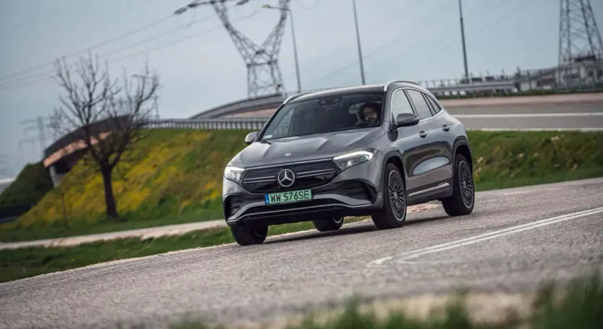 Szary Mercedes-Benz EQA jedzie po drodze, w tle linie energetyczne i drzewo.