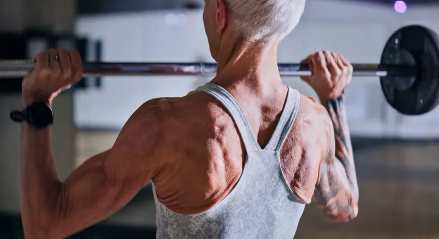 Stark ryggmuskulatur hos en kvinna som lyfter skivstång. Hon har tatuerade armar och en träningsklocka. För att bygga muskler är det viktigt med tillräckligt med gram protein per kg kroppsvikt.