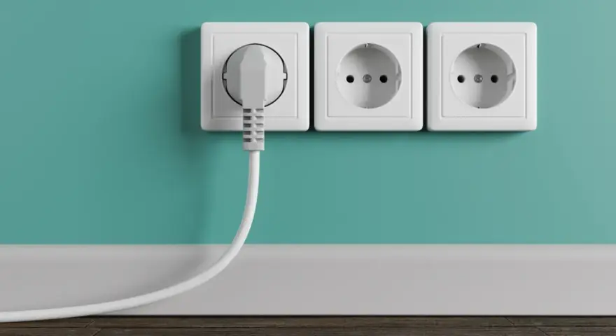 Tres enchufes blancos en una pared turquesa, uno con un enchufe gris conectado. La altura de los enchufes en la cocina es estándar.